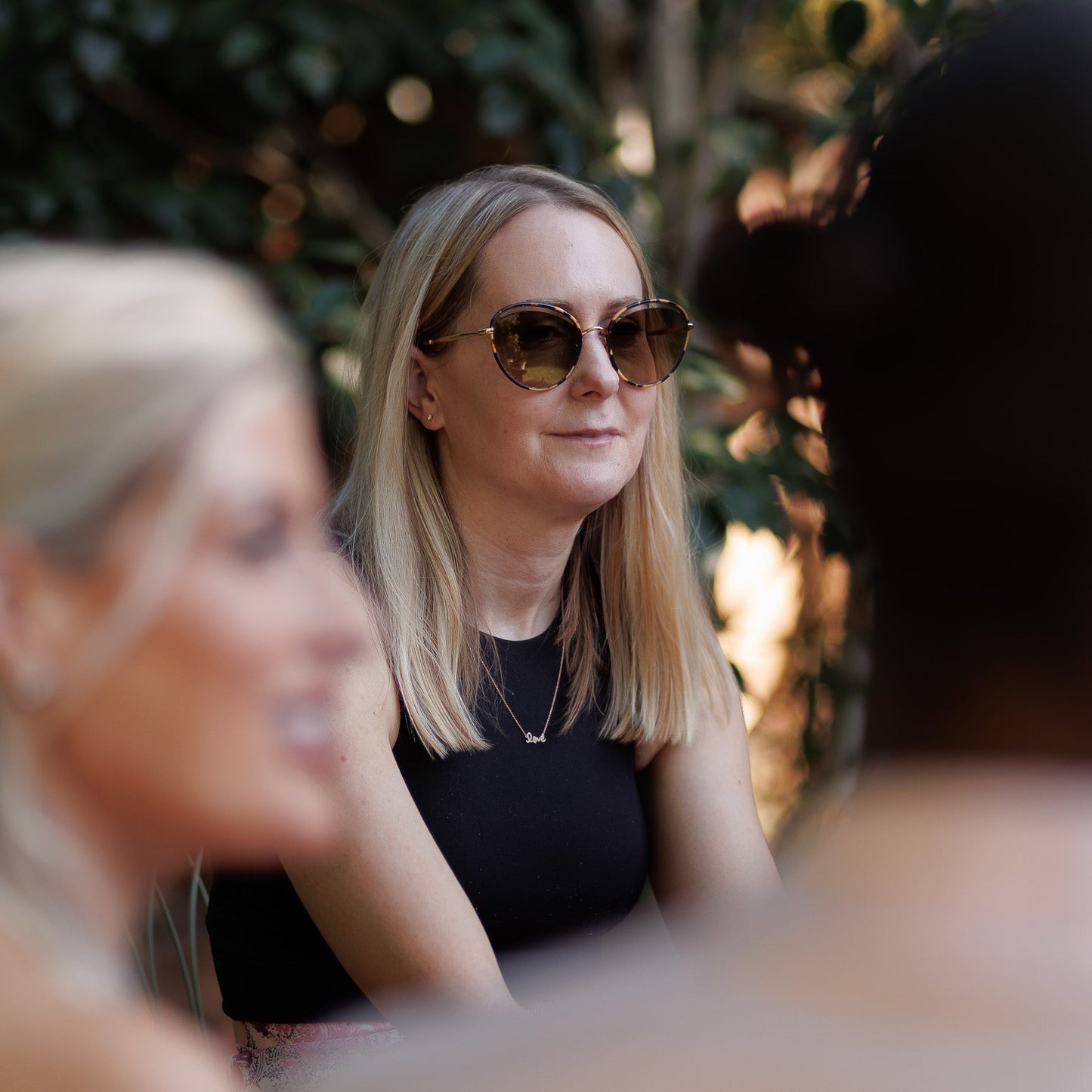 A woman with blonde hair and brown aviator-style sunglasses wearing a black sleeveless top, captured in a warm, outdoor social setting with blurred foreground figures.