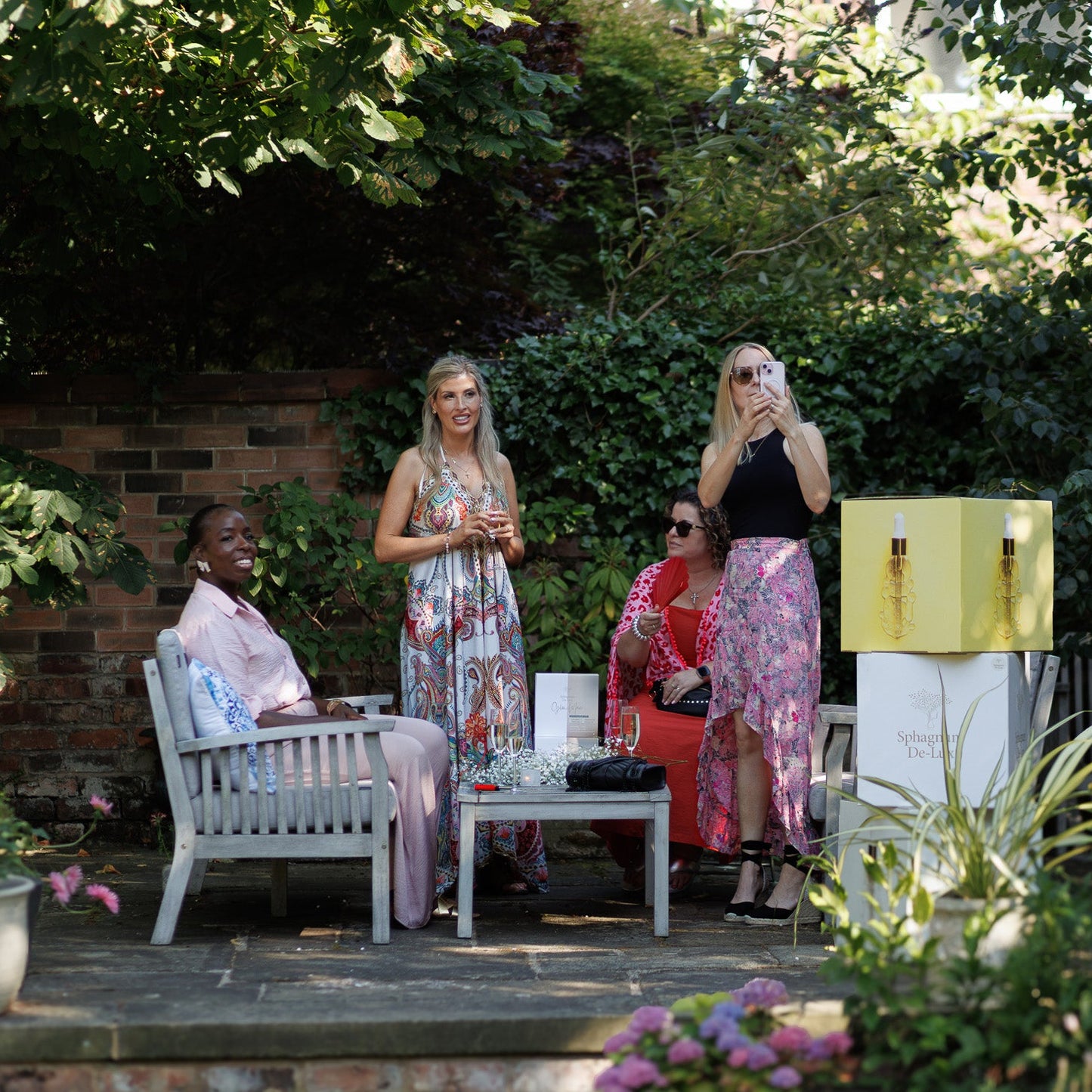 Three women are gathered outdoors in a garden patio setting. One woman sits on a white chair next to a small table, while the other two stand nearby.