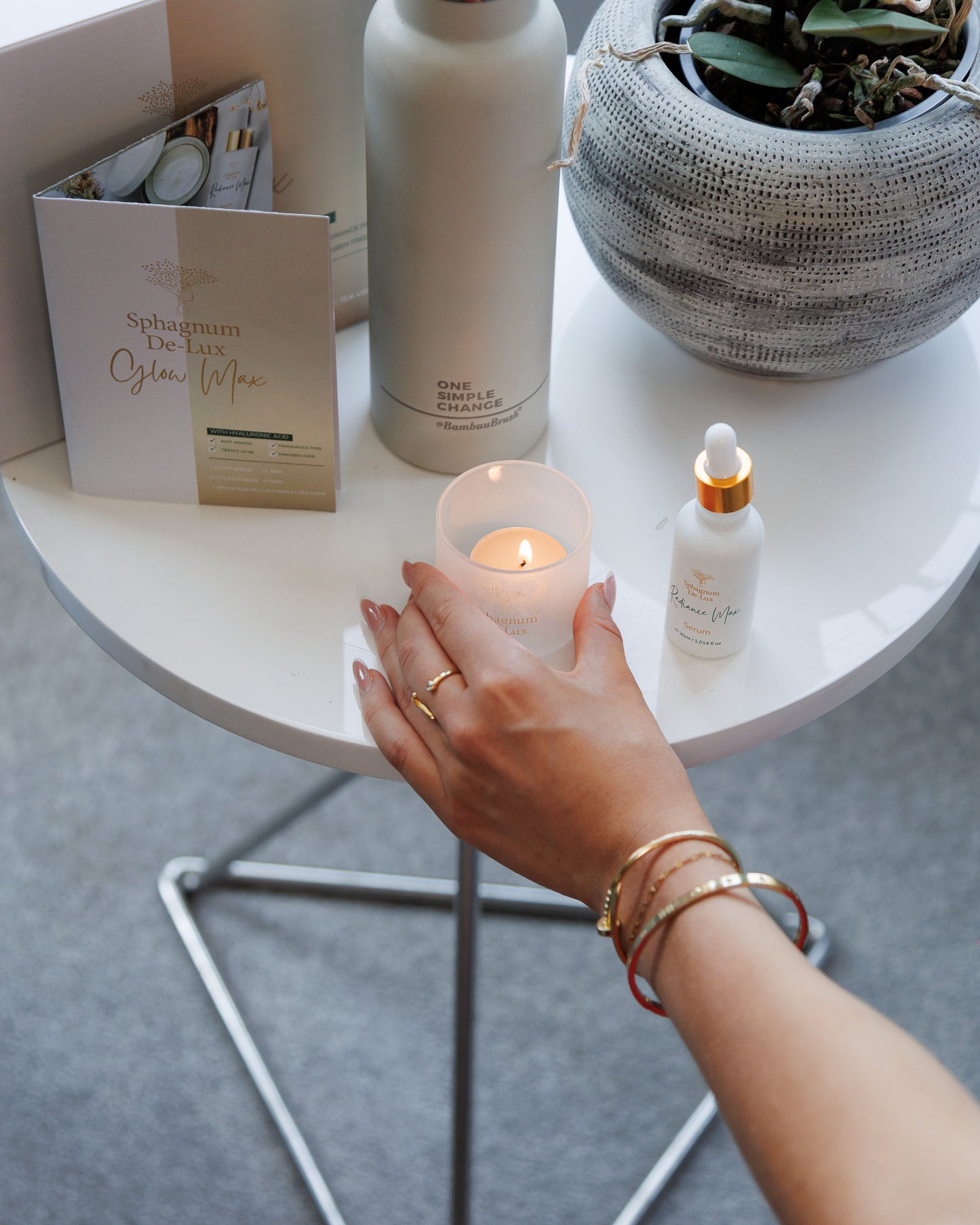 A lifestyle shot of a skincare setup on a white round table, featuring a hand holding a lit candle, a white bottle of Sphagnum Dampener, a small dropper bottle, and a greeting card.