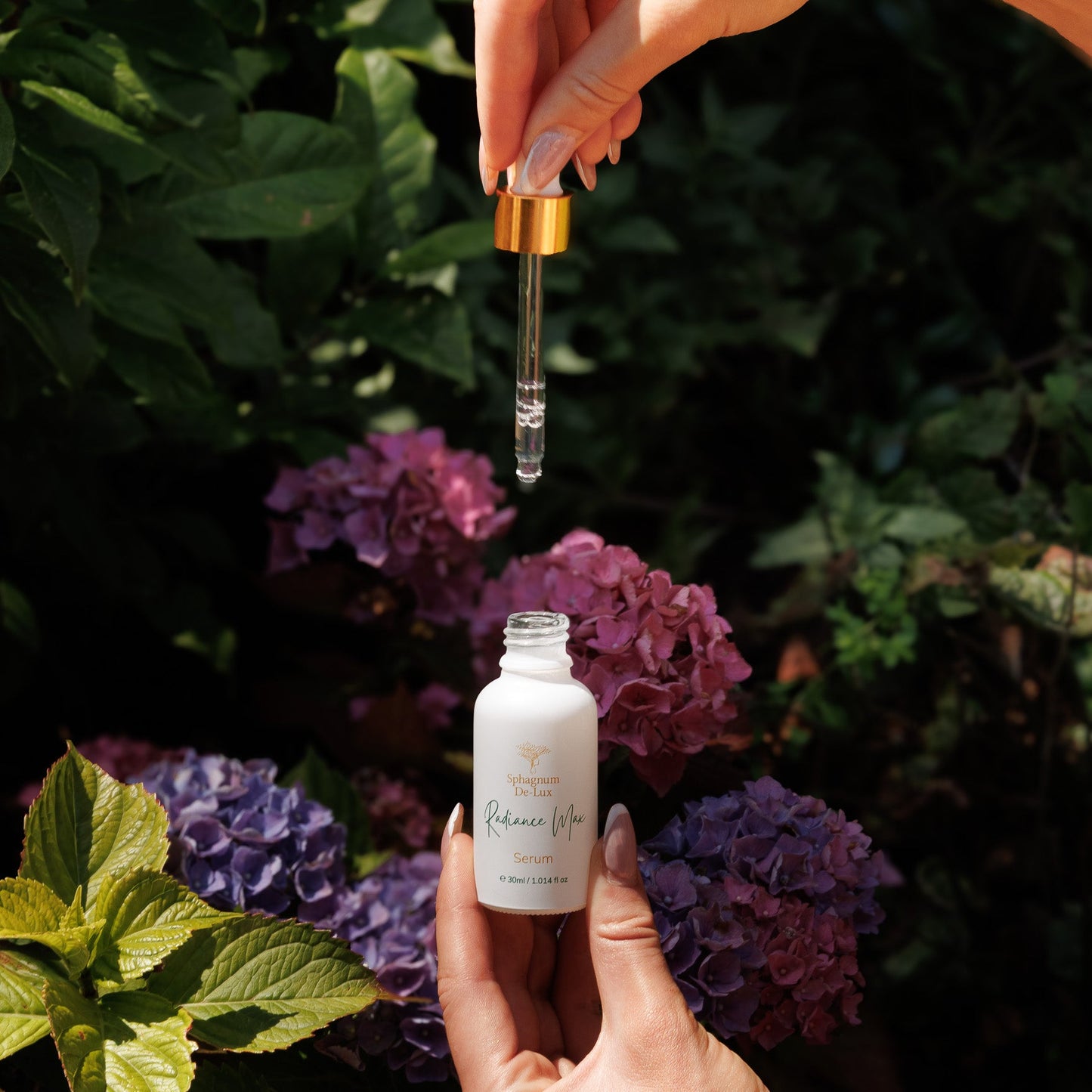 A hand holds a small white bottle labeled 'Calming Face' while another hand drops a clear liquid dropper above it, set against a background of green leaves and purple hydrangeas.